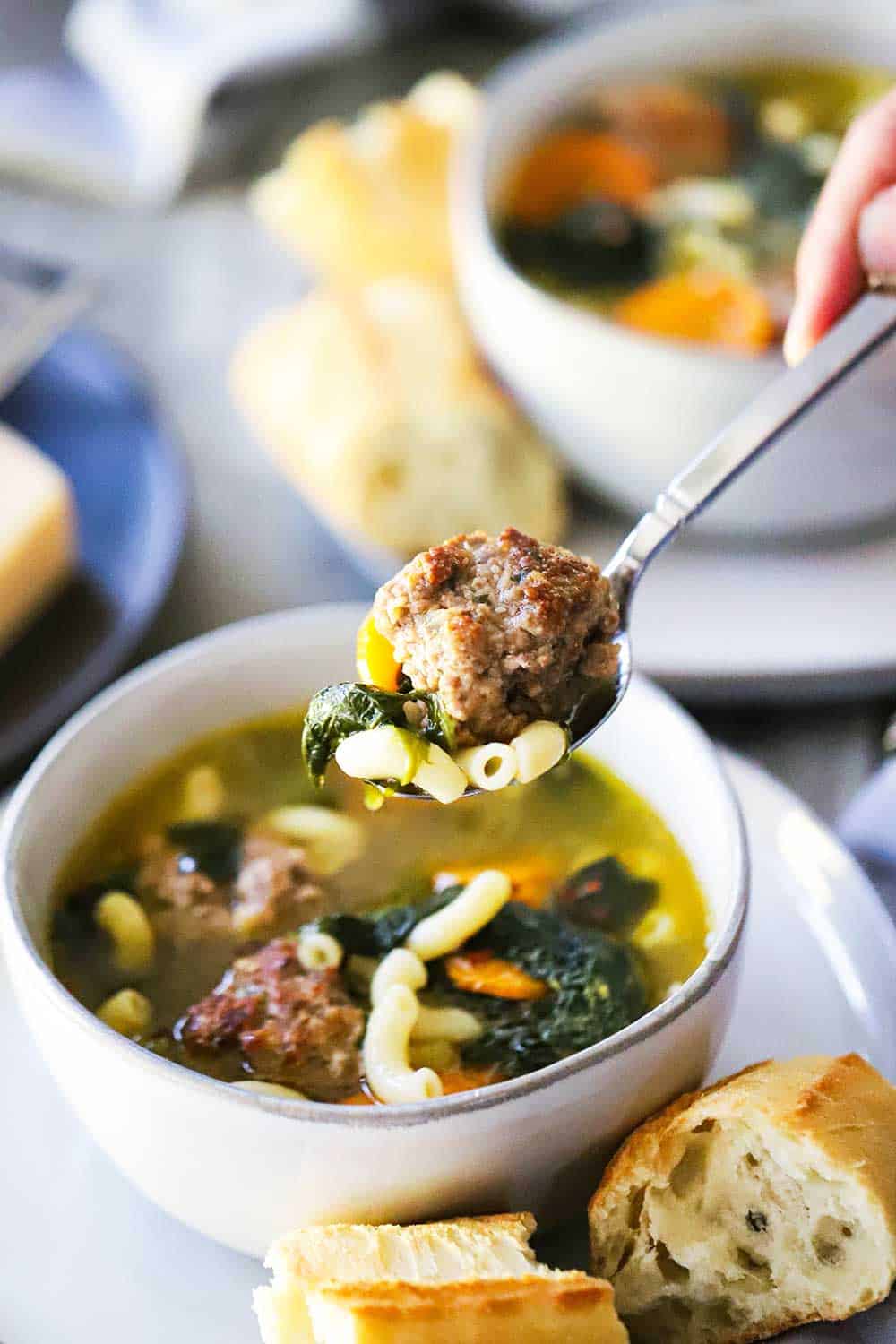 A hand holding a soup spoon with a meatball, some pasta, and spinach on it and over a bowl of the soup.