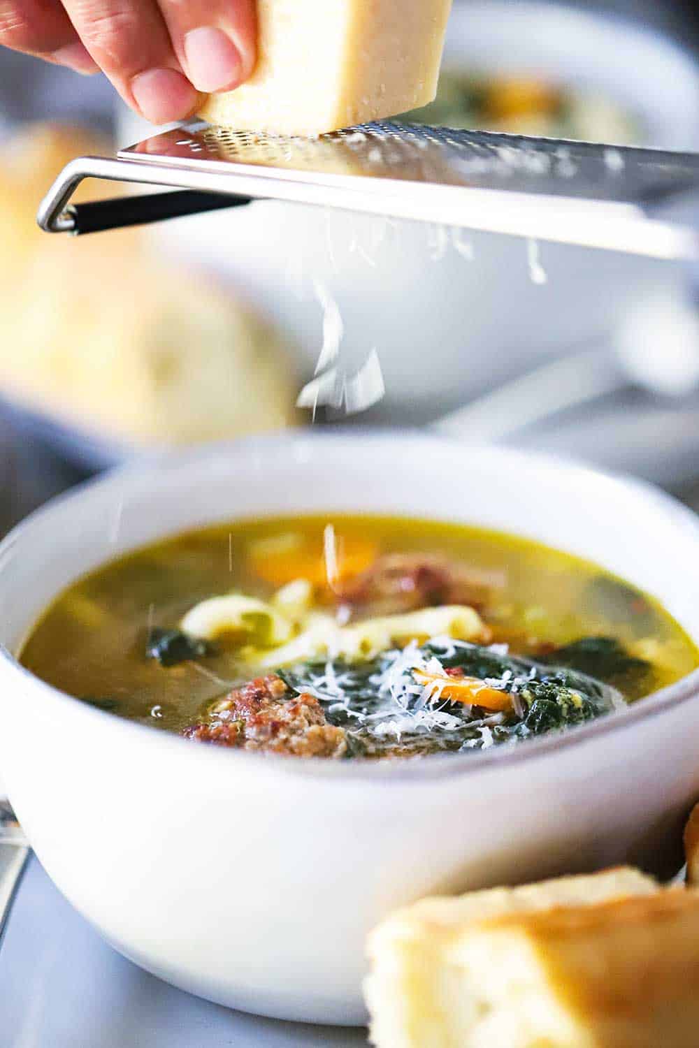 A hand scraping a block of Parmesan cheese across a microplane over a bowl of Italian wedding soup.