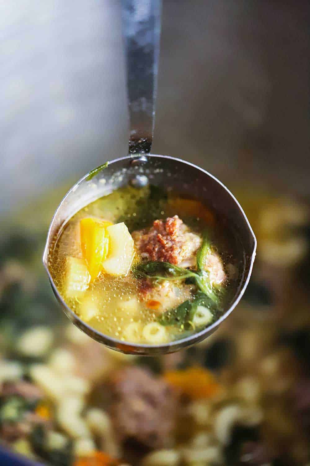 A large ladle filled with soup containing a meatball, pasta, spinach, and chicken stock.