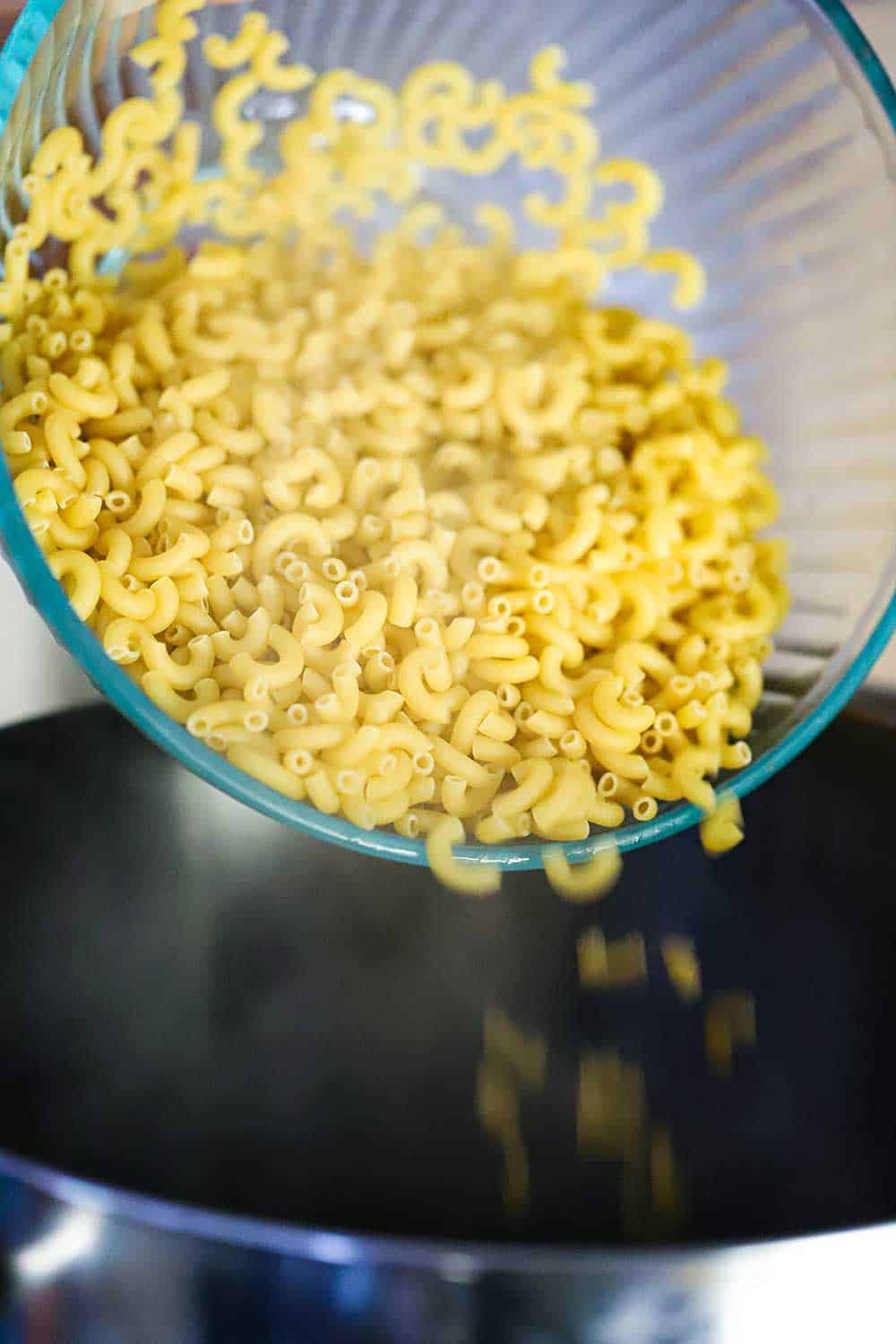 A glass bowl full of uncooked elbow pasta being dropped into a large stock pan.