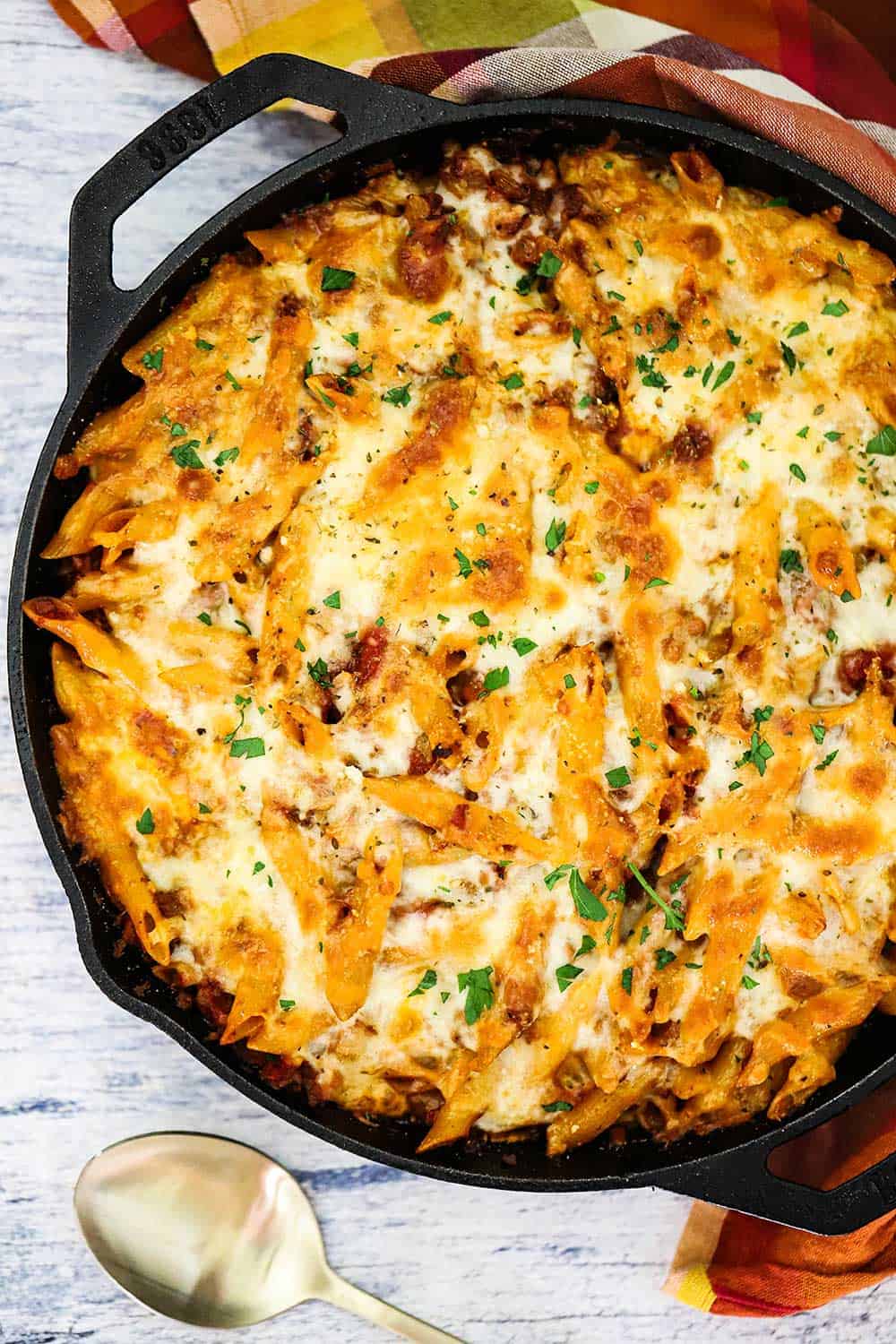 A large cast-iron skillet filled a vegetarian pasta bake next to a gold spoon. 