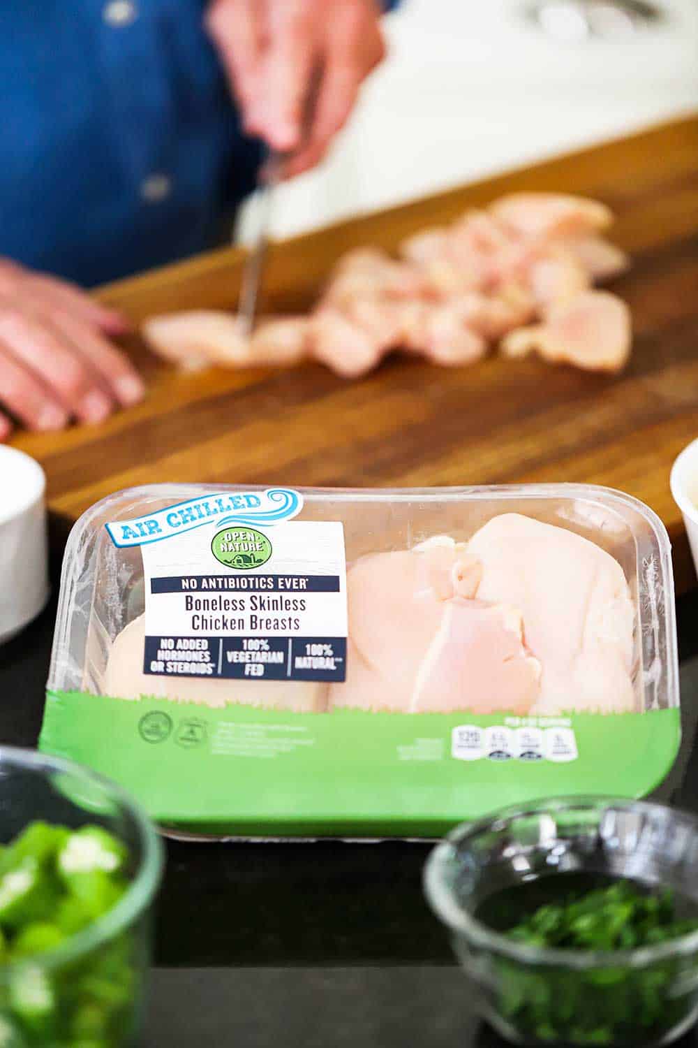 A person slice pieces of a chicken breast on a cutting board with a package of chicken breast in front of it.