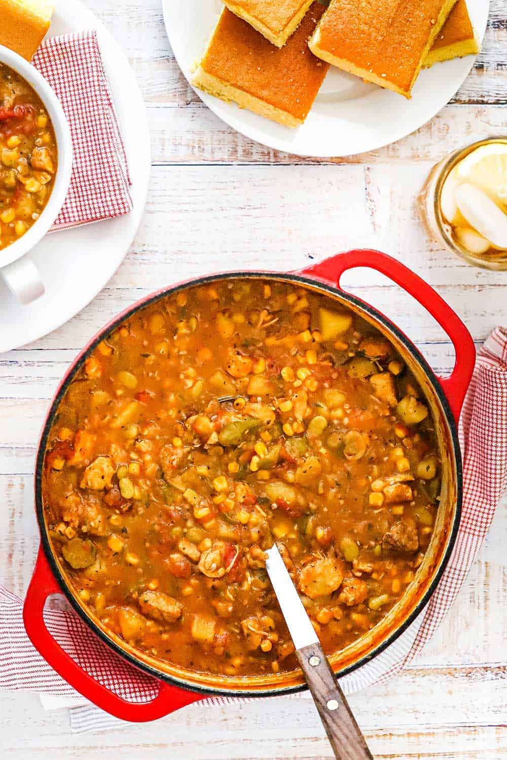 A large red circular Dutch oven filled with Brunswick stew next to a plate of yellow cornbread pieces. 