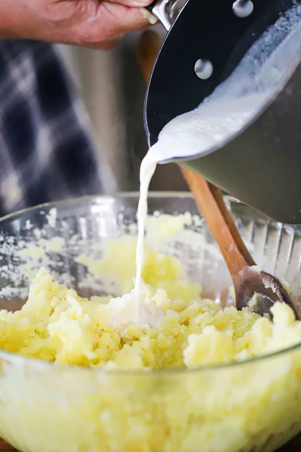 Warm cream being poured into a large bowl filled with mashed potatoes. 