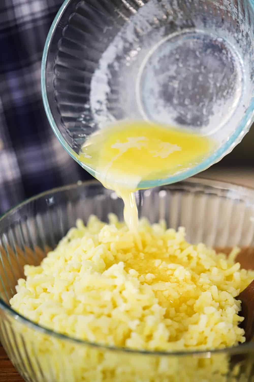 Melted butter being poured from a small glass bowl into another large bowl filled with cooked gold potatoes. 
