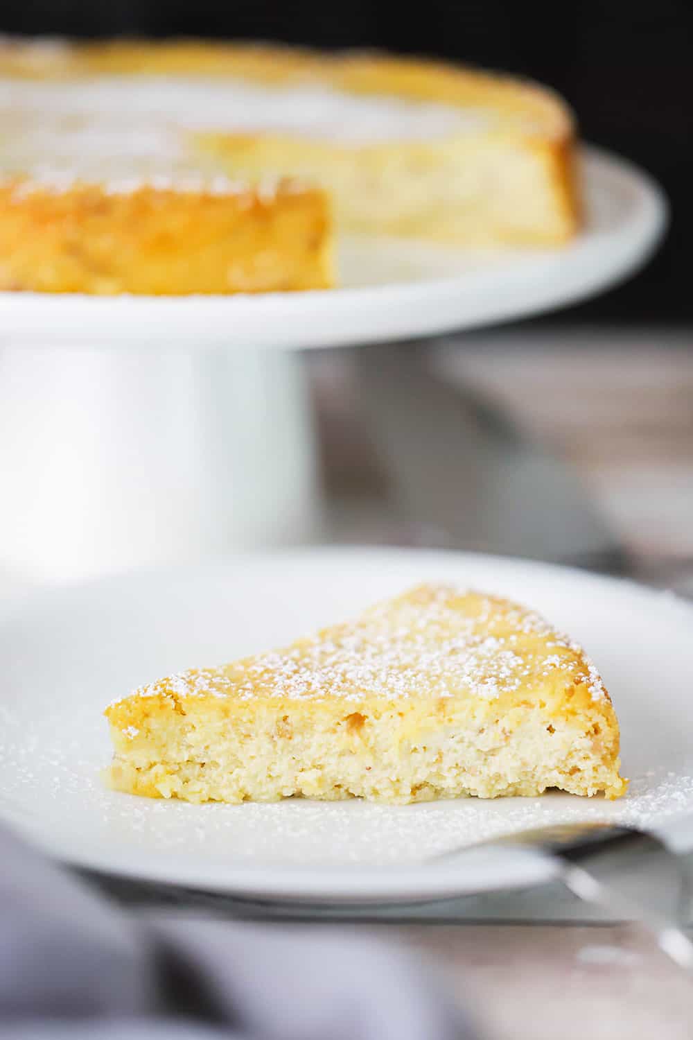 A slice of lemon ricotta cheesecake on a plate next to cake stand holding the cake. 