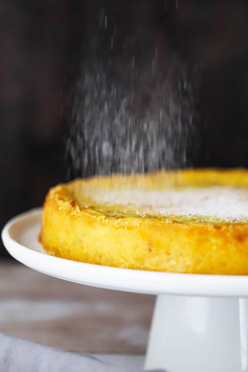 Powdered sugar sprinkling over the top of Italian ricotta cheesecake on a cake stand. 