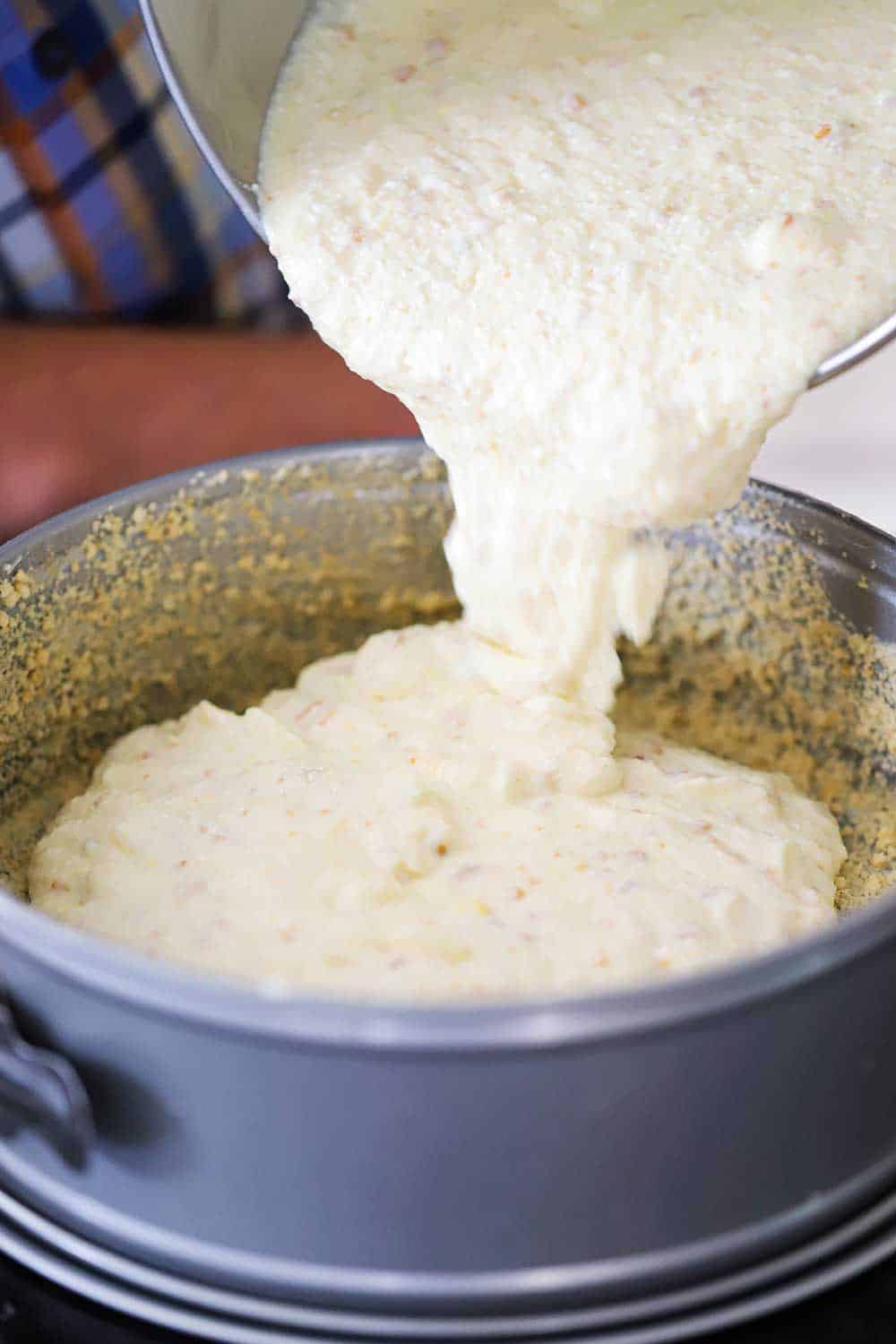 Ricotta cheesecake batter being poured into a springform pan lined with butter and cookie crumbs. 