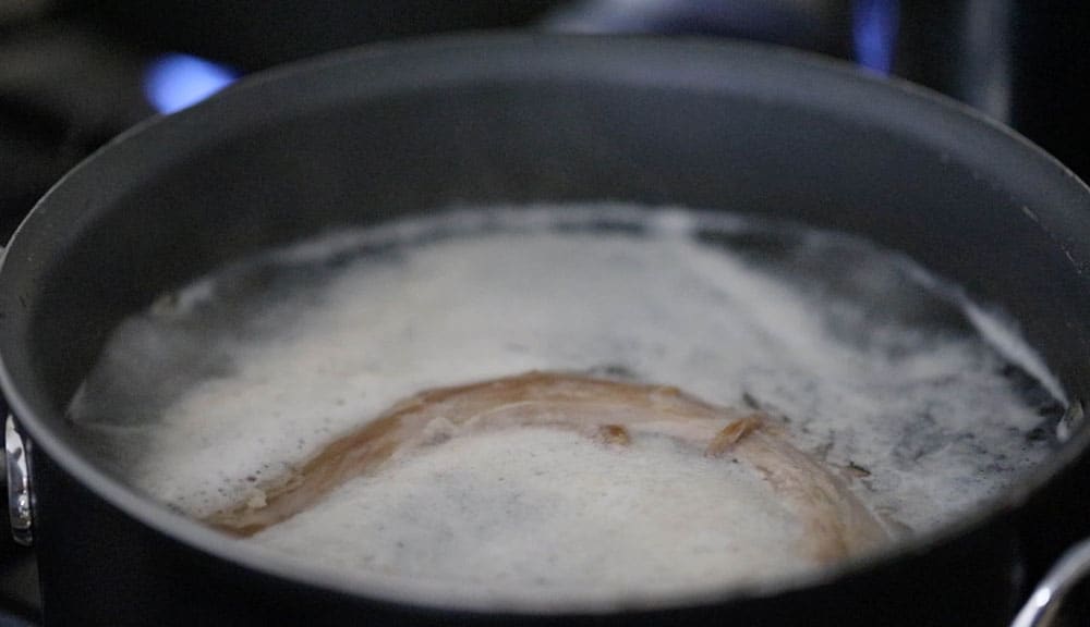 A turkey neck in a pot that is simmering in water with impurities that have floated to the surface.