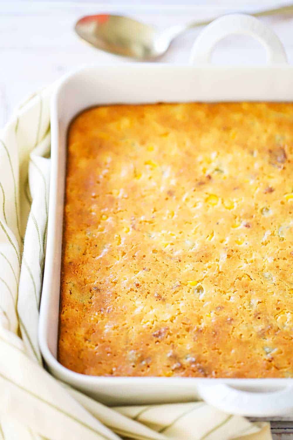 A fully cooked corn casserole in a square white baking dish next to a gold serving spoon. 