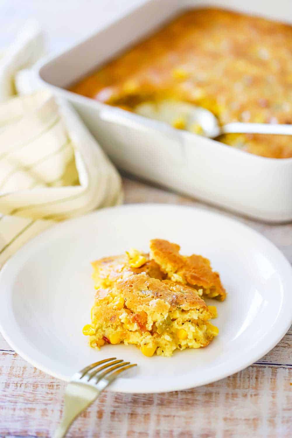 An individual serving of corn casserole on a small plate next to a baking dish filled with the casserole. 