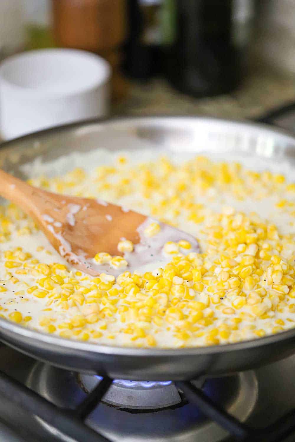 A large silver skillet on the stove filled with homemade creamed corn with a wooden spoon in it. 