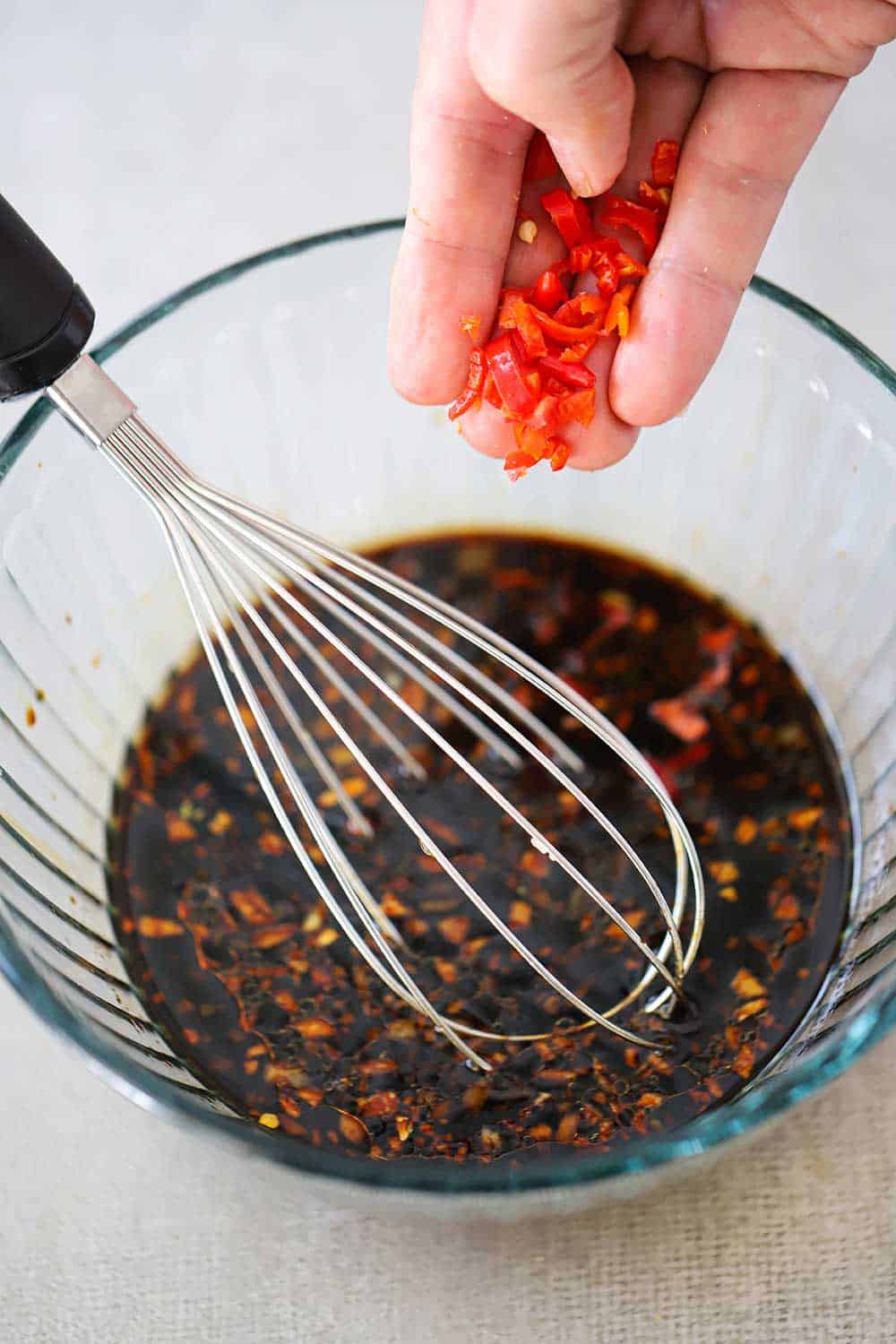 A hand dropping chopped red peppers into a glass bowl of ginger soy dipping sauce.