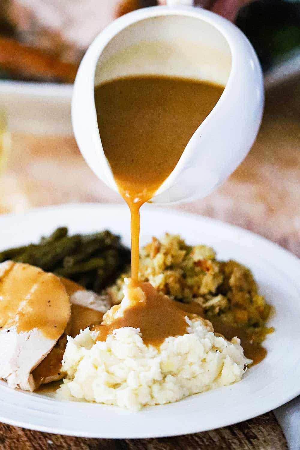 A white gravy boat pouring turkey gravy over a plate of mashed potatoes, dressing, turkey, and green beans.