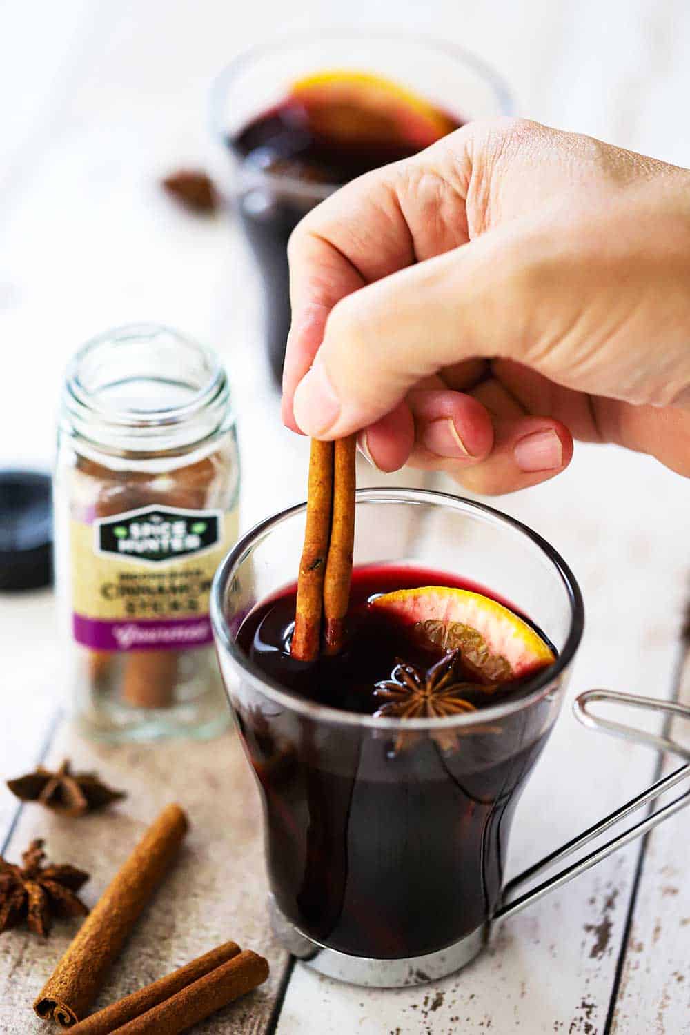 A hand placing a cinnamon stick into a clear glass filled with mulled wine.