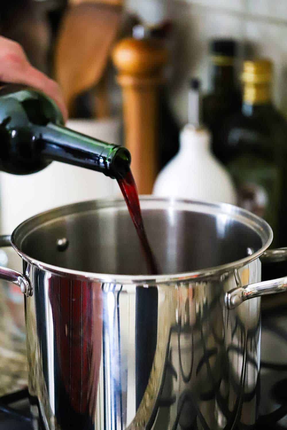 A hand pouring red wine from a bottle into a large silver pot on the stove.