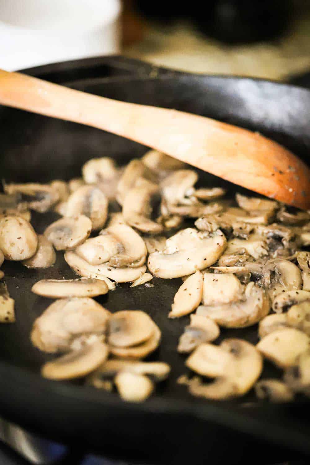 A black cast-iron skillet with sliced mushrooms being sautéd in it, with a wooden spoon in the pan, also.