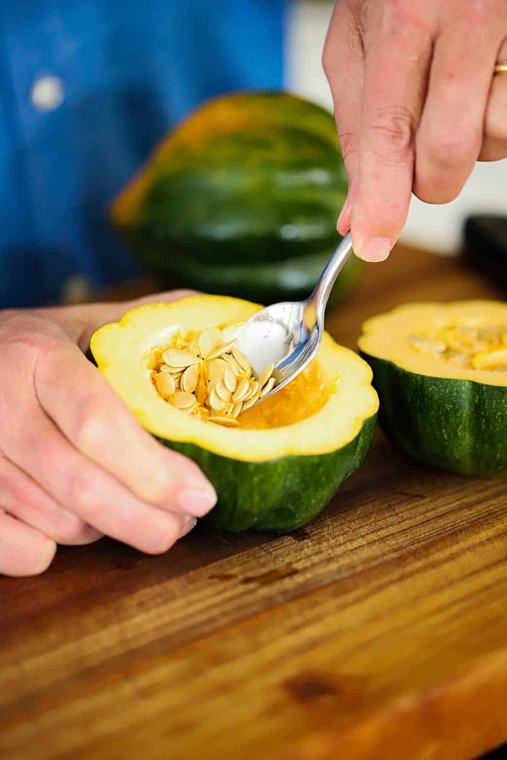 A person using a spoon to remove seeds from the center of an acorn squash that has been cut in half. 