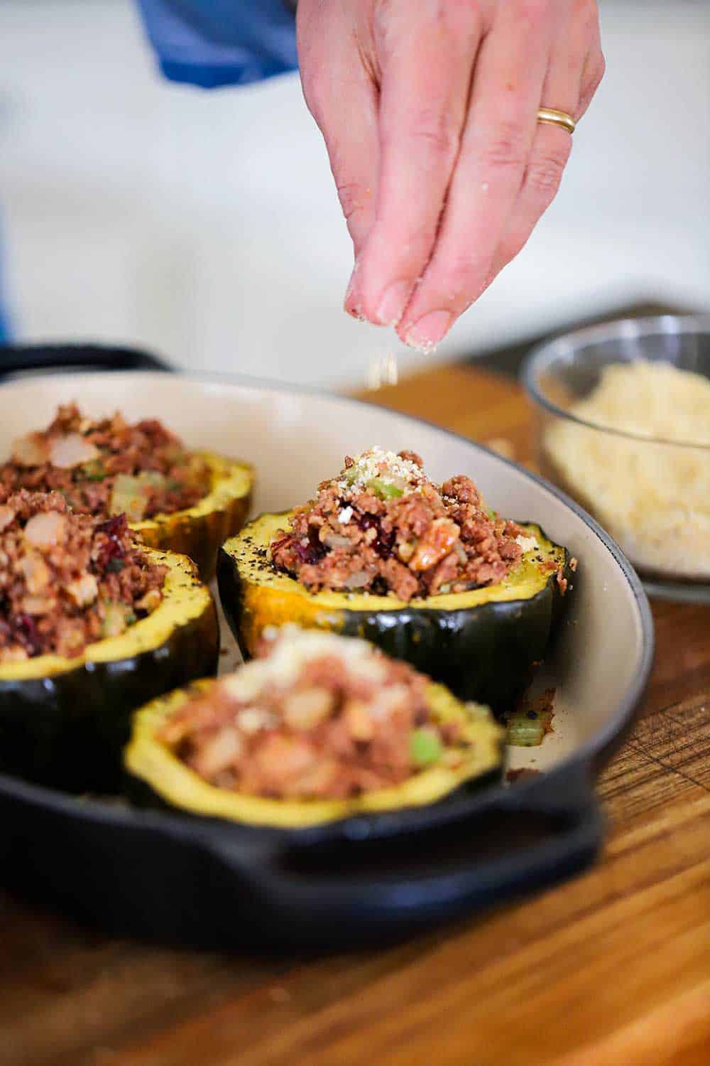 A hand sprinkling bread crumbs onto of stuffed acorn squash in a baking dish. 