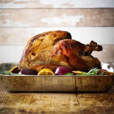 A roasted turkey sitting in a steel roasting pan surrounded by lemons, onions, and herbs.