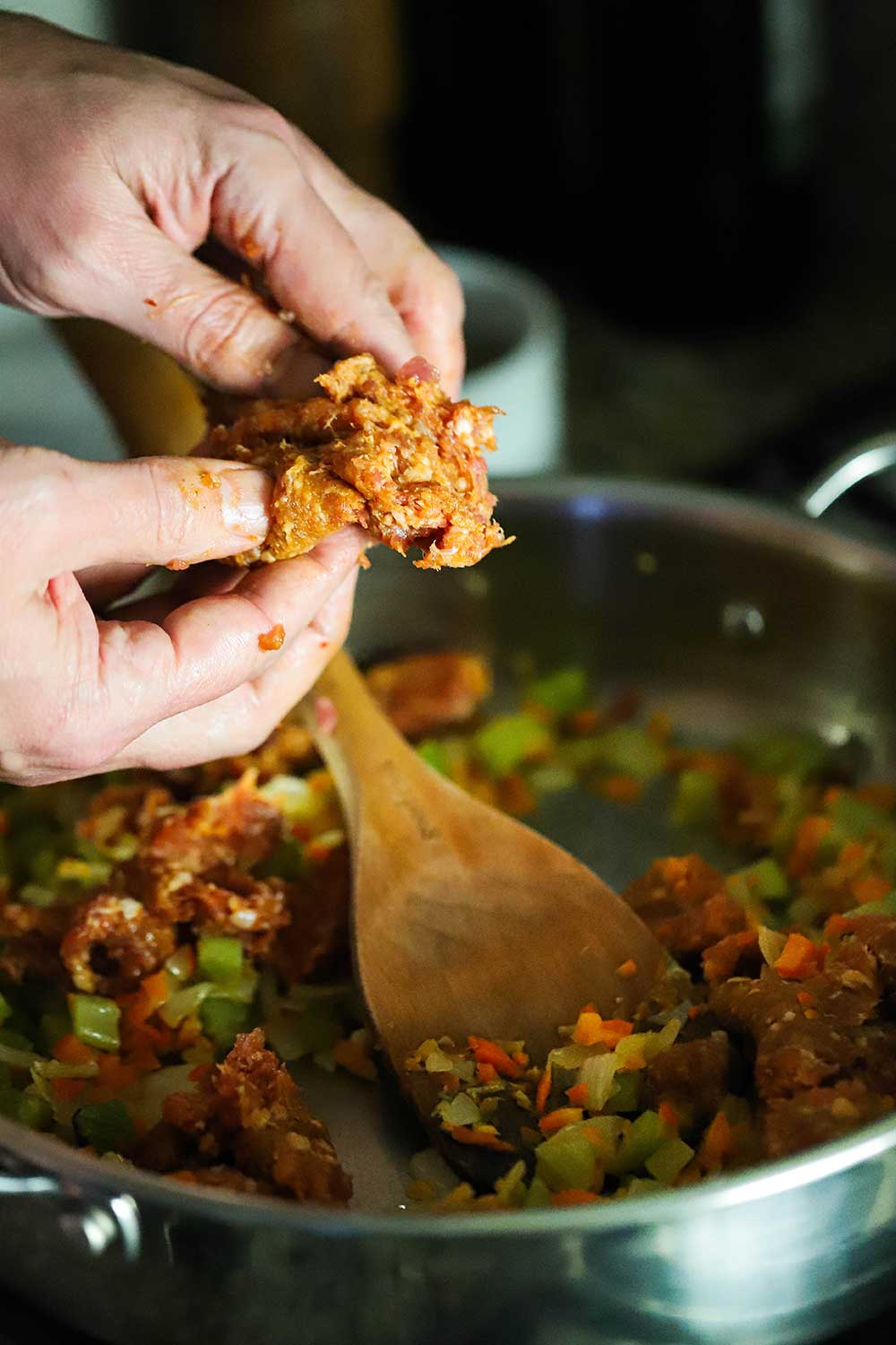 Two hands crumbling uncooked Italian sausage into a skillet filled with sautéd vegetables.