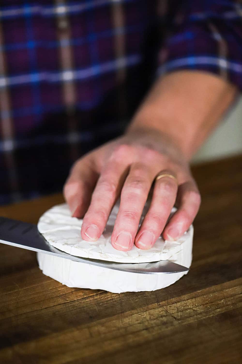 A person using a chef's knife to slice the top layer of rind from a wheel of brie. 