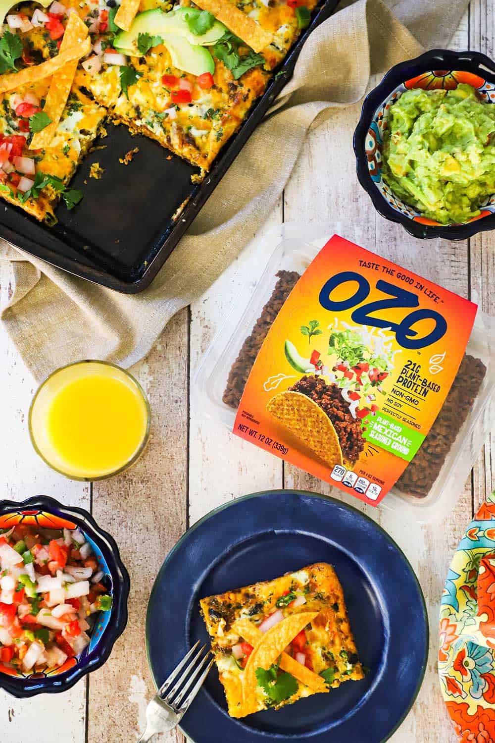 A blue plate with a slice of Mexican frittata on it sitting next to a package of OZO plant-based Mexican ground and a glass of orange juice. 