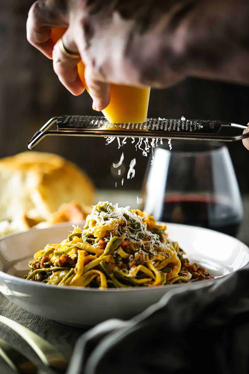 A hand grating a block of Parmesan cheese over white bowl filled with fettuccine with creamy sausage sauce.