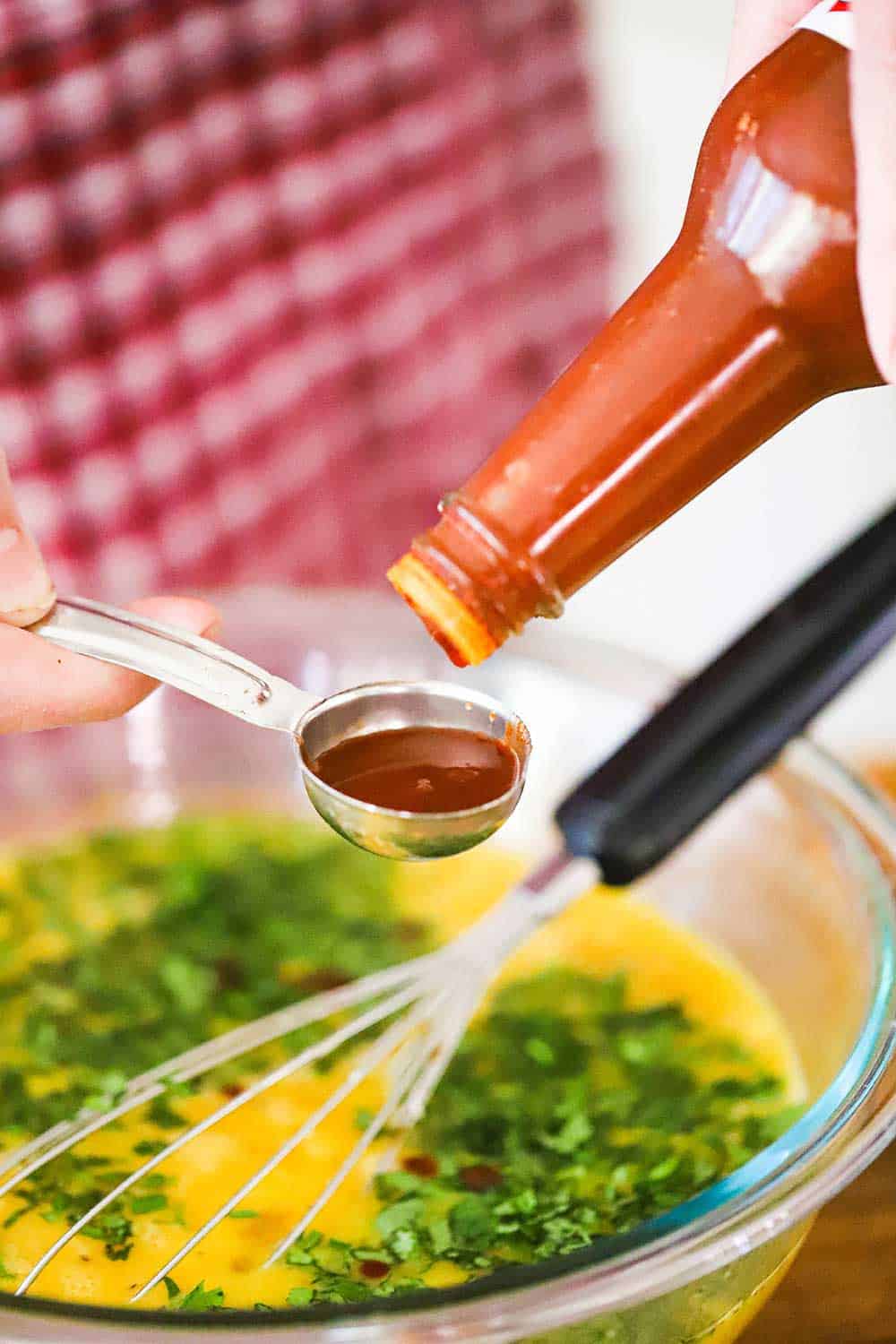 A person pouring hot sauce into a tablespoon over a large glass bowl filled with eggs and chopped cilantro. 