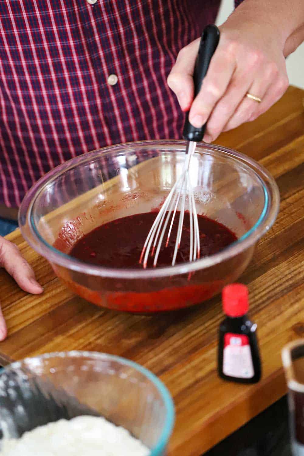 A person using a whisk to mix together red food dye into melted chocolate in a glass bowl. 