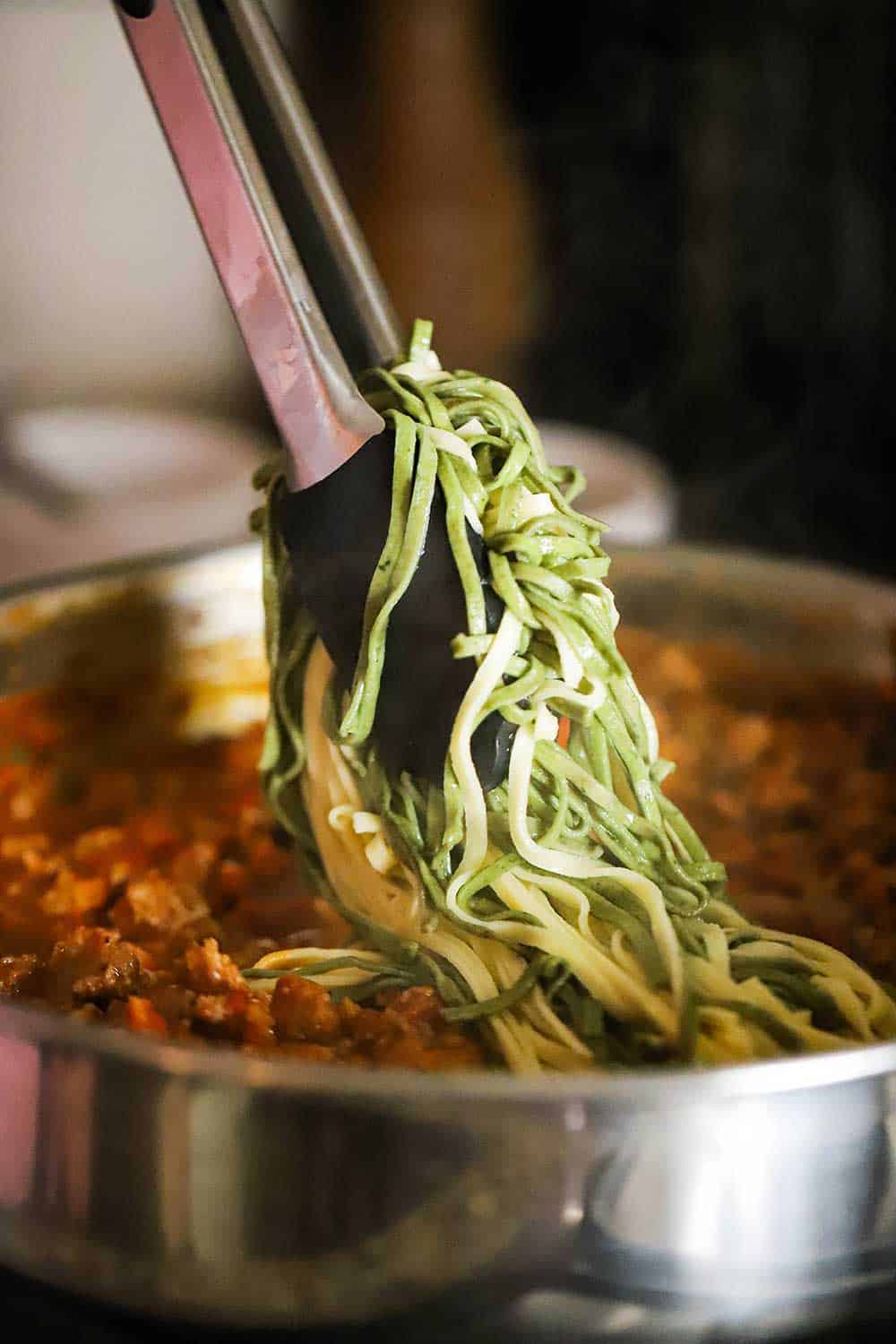 A pair of tongs lowering spinach and egg pasta into a skillet of creamy sausage sauce.