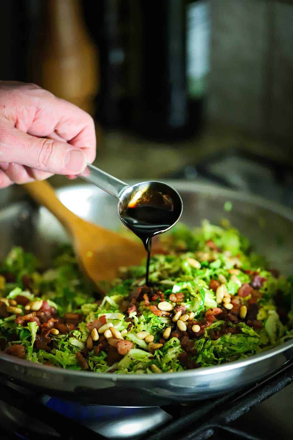 A hand pouring a balsamic glaze from a tablespoon into a skillet of cooked shaved Brussels sprouts and pancetta. 