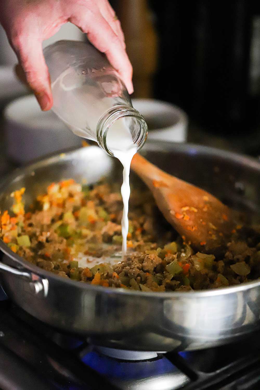 A hand pouring cream from a small milk jug into a skillet filled with cooked sausage and vegetables.