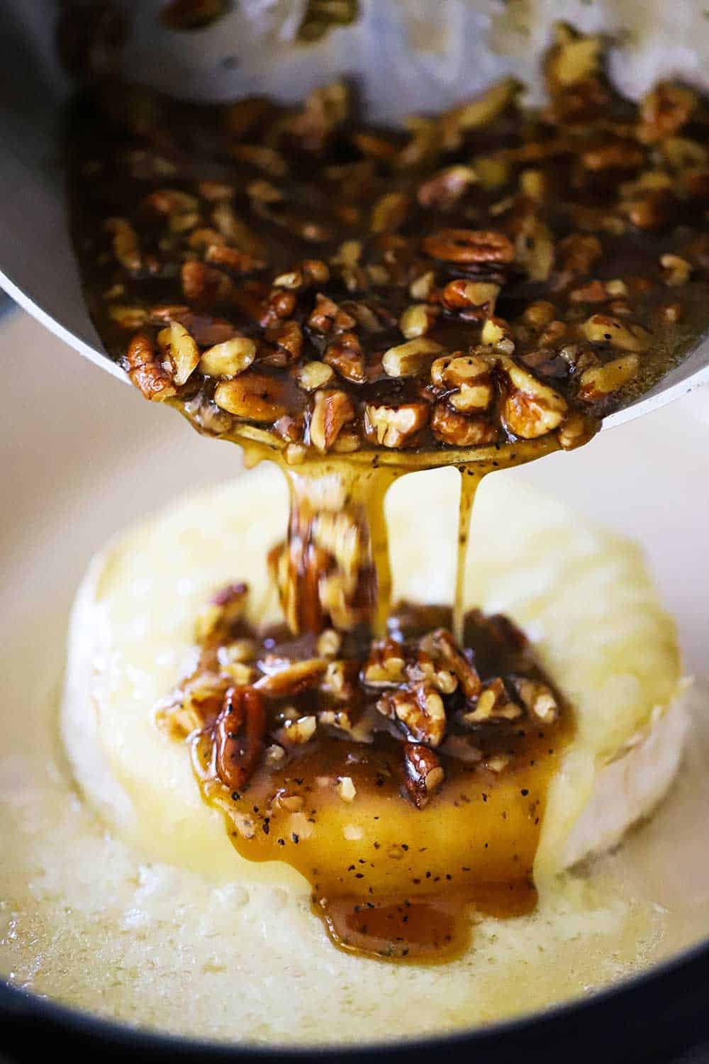 A medium-sized saucepan being used to pour a pecan praline sauce over baked brie. 