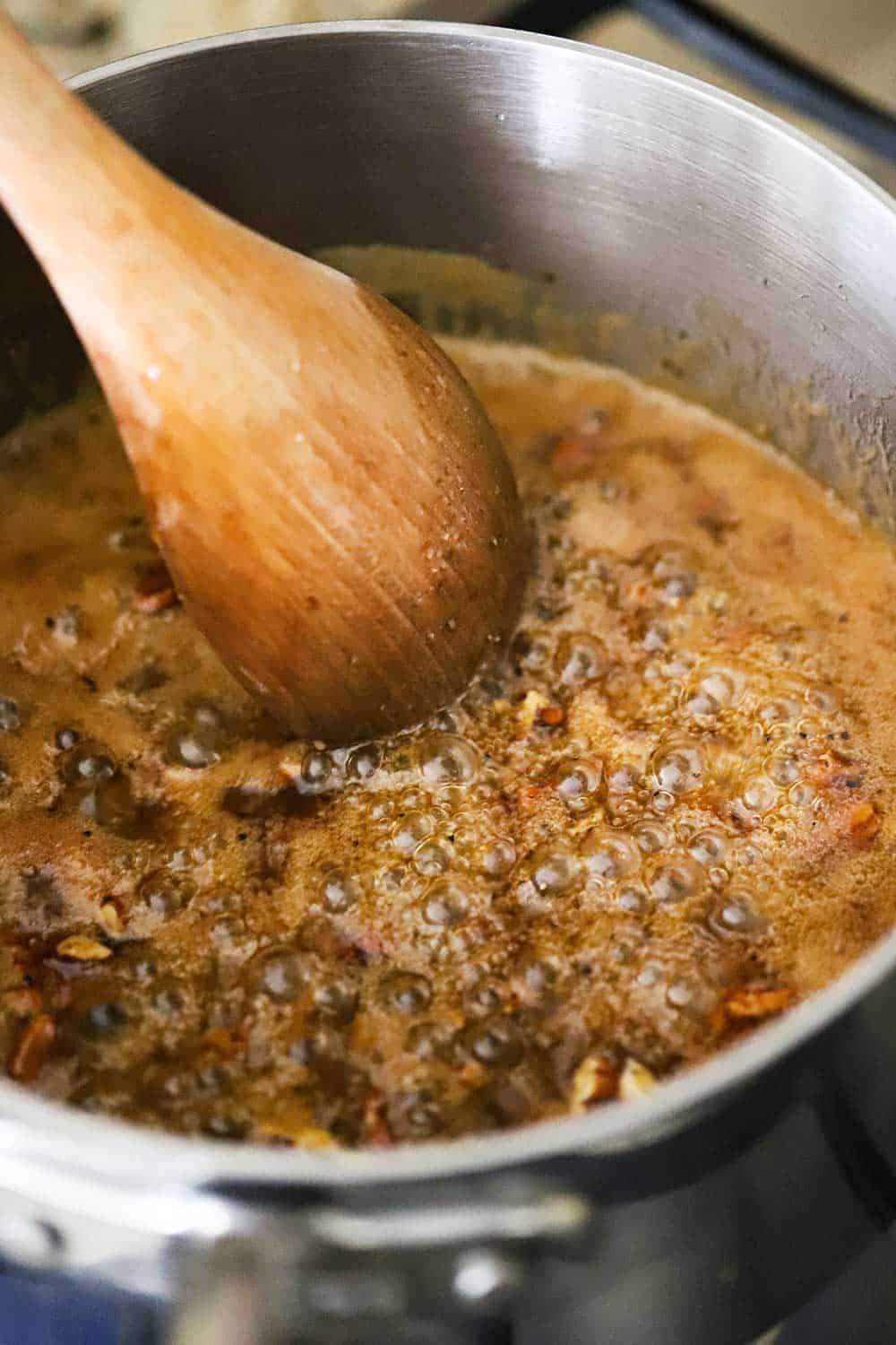 A stainless steel saucepan filled with simmering praline sauce with a wooden spoon in it. 