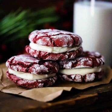 Three red velvet sandwich cookies stacked on a piece of brown paper next to a glass of milk.