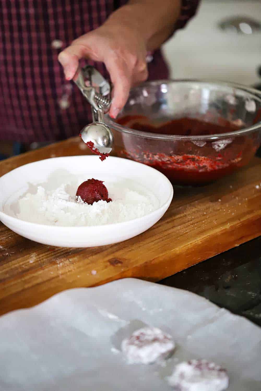 A person using a trigger handle ice cream scooper to drop red velvet cookie dough into a bowl filled with powdered sugar. 