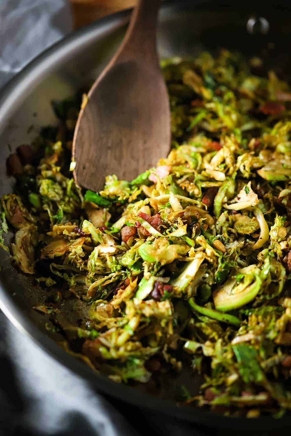 A large steel skillet filled with cooked shaved Brussels sprouts with pancetta and balsamic and a wooden spoon in the middle of the pan. 