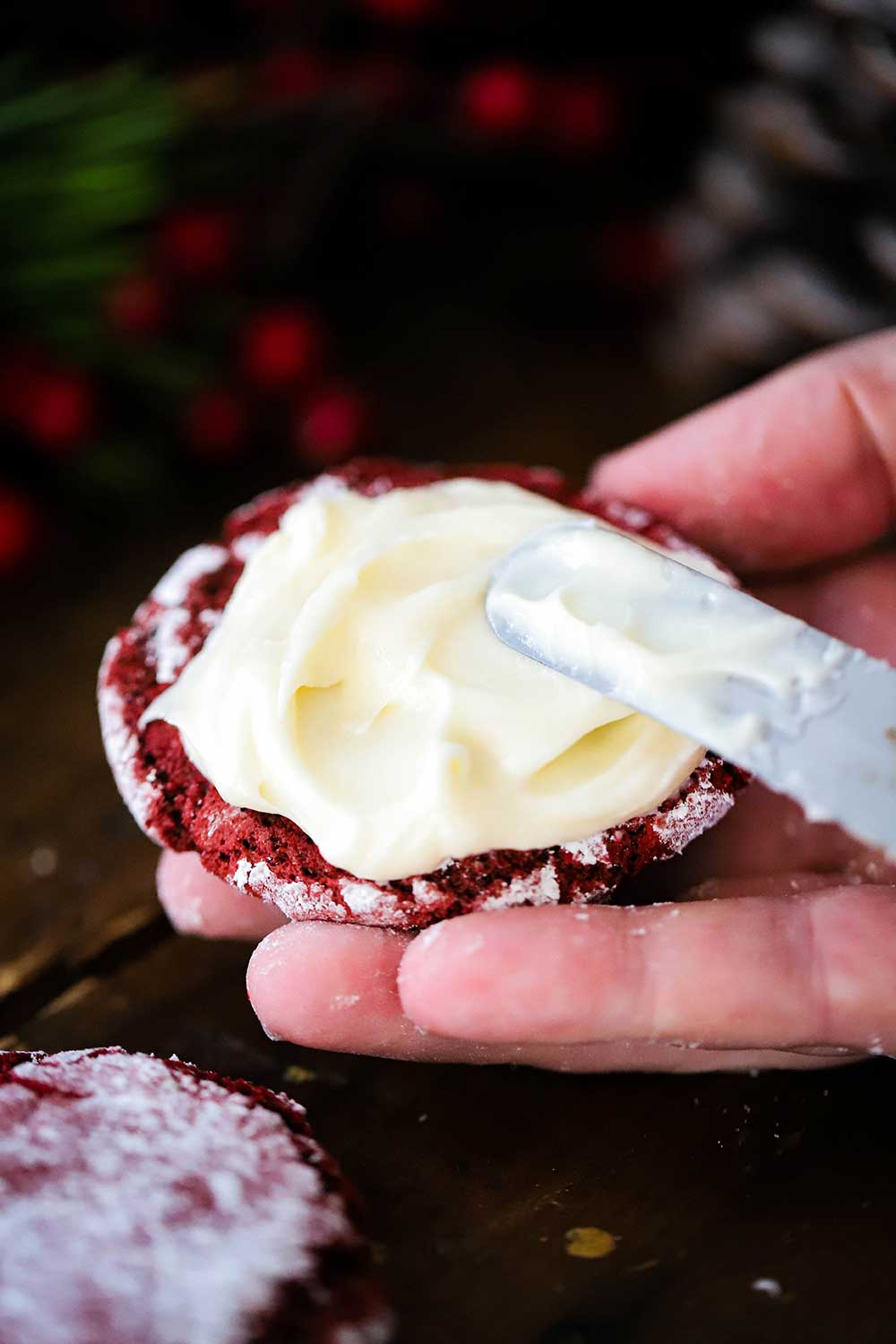 An off-set spatula spreading cream cheese frosting on the flat side of a red velvet cookie. 