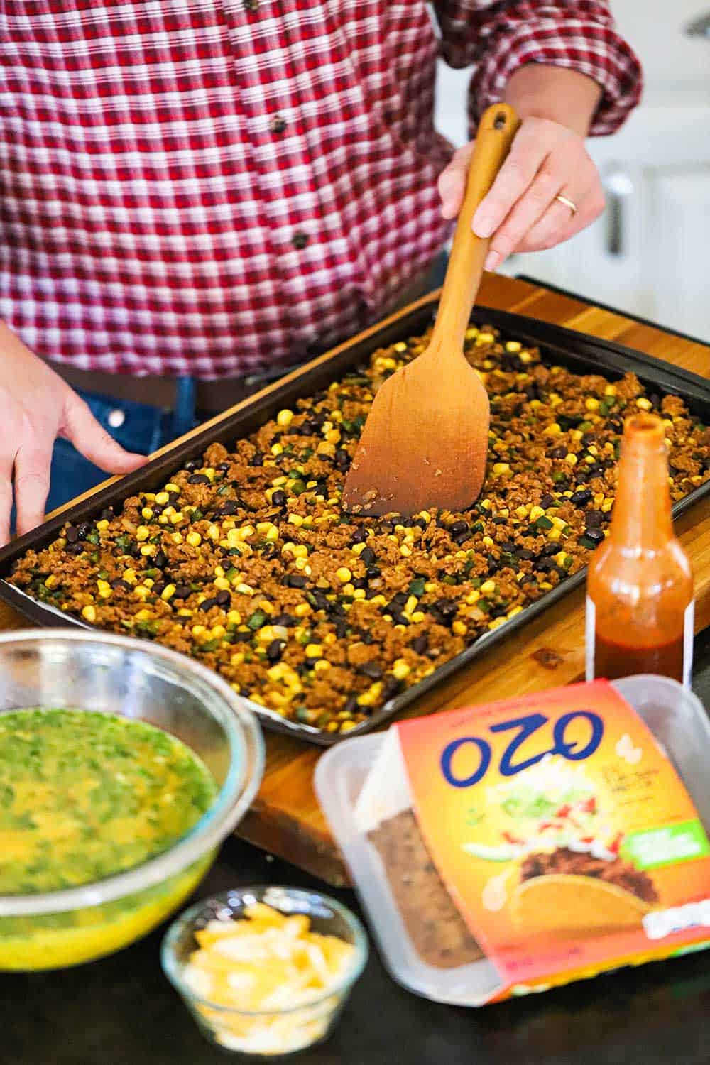 A person using a wooden spatula to spread a plant-based Mexican mixture over the base of a rimmed baking sheet pan. 