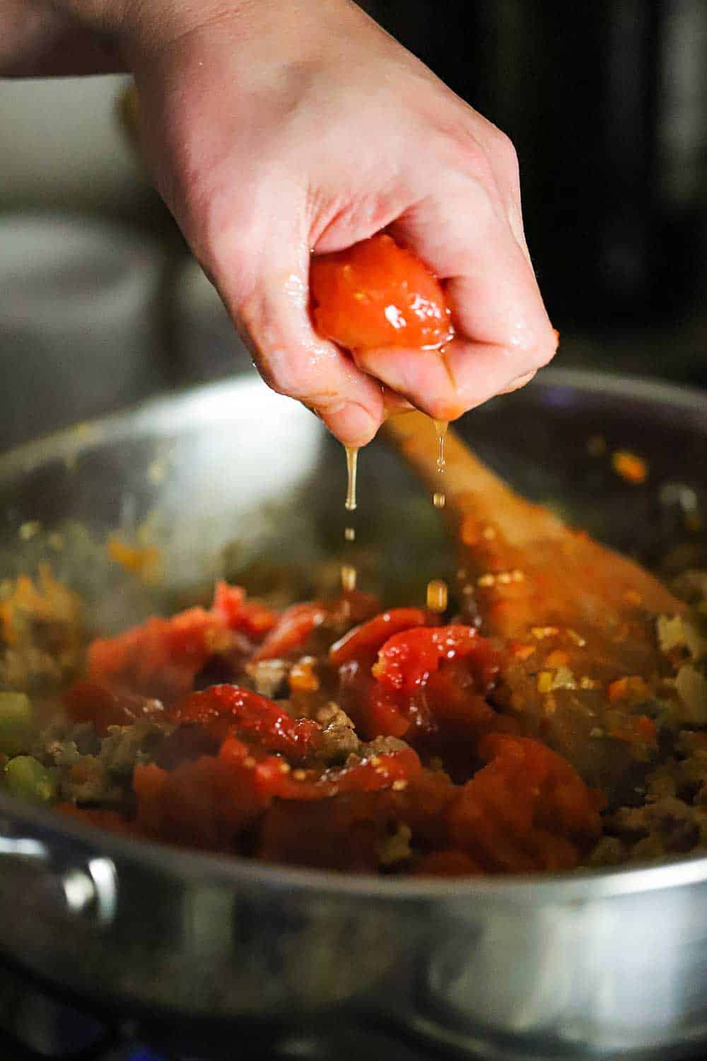 A hand squeezing a whole tomato into a skillet of sautéd vegetables.
