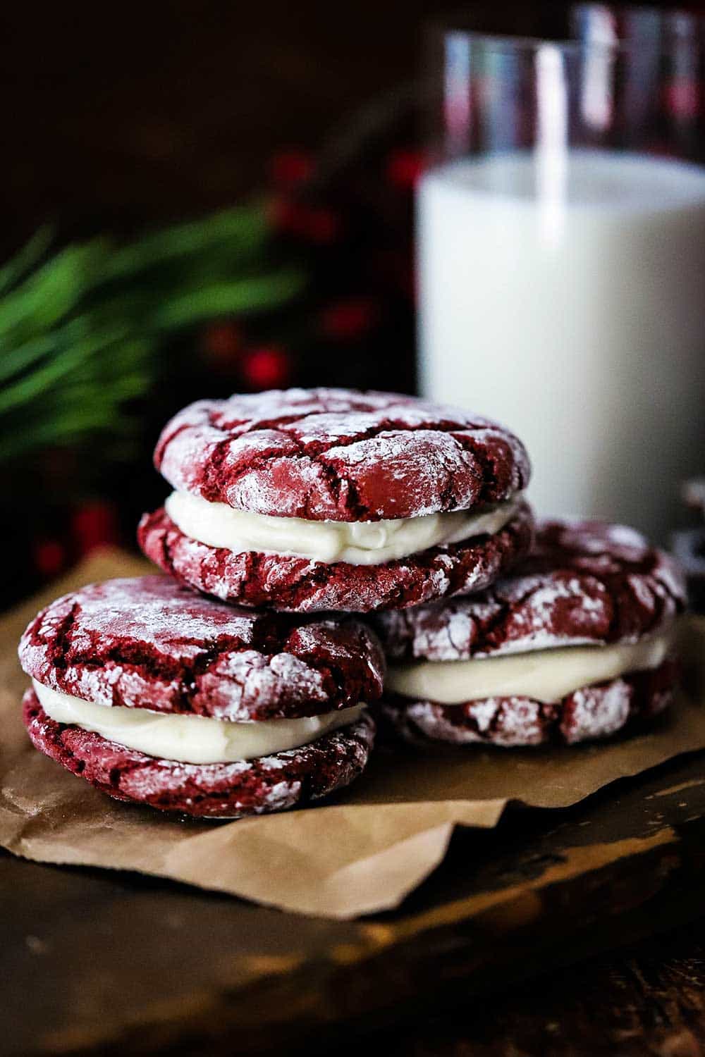 Three red velvet sandwich cookies stacked on a piece of brown paper next to a glass of milk. 
