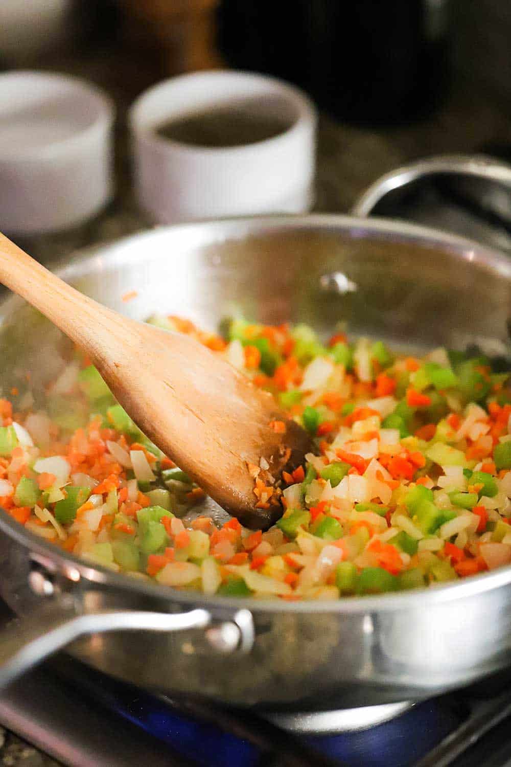 A large stainless steel skillet filled with sautéd mirepoix with a wooden spoon in the middle of it.