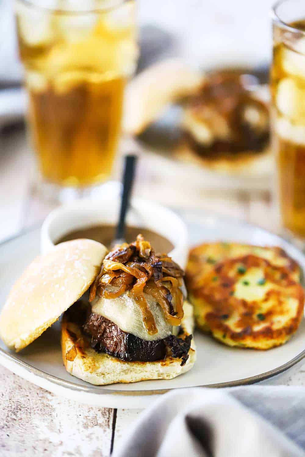 A white plate filled with a pot roast slider topped with caramelized onions sitting next to two potato cakes, with a small bowl of gravy next to it, as well. 