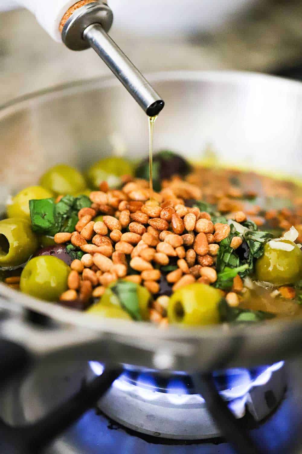 Olive oil being drizzled into a small saucepan filled with toasted pine nuts, olives, garlic, and white wine over a burner on a stove.