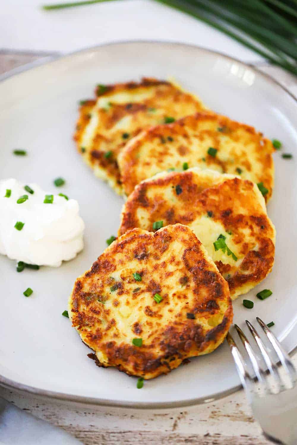 5 leftover mashed potato cakes layered on a plate with a dollop of sour cream nearby and sprinkled with chopped chives. 