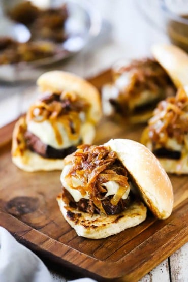 Four pot roast sliders each topped with melted Swiss cheese and caramelized onions all on a small cutting board.