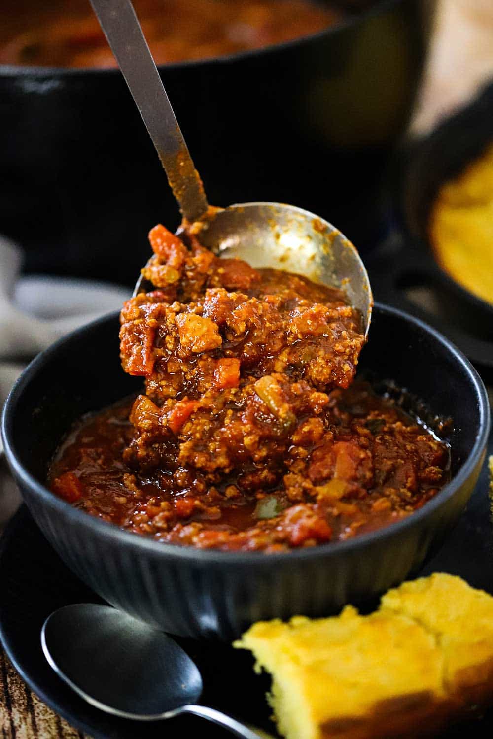 A helping of turkey chili being ladled into a dark blue bowl next to a piece of cornbread.