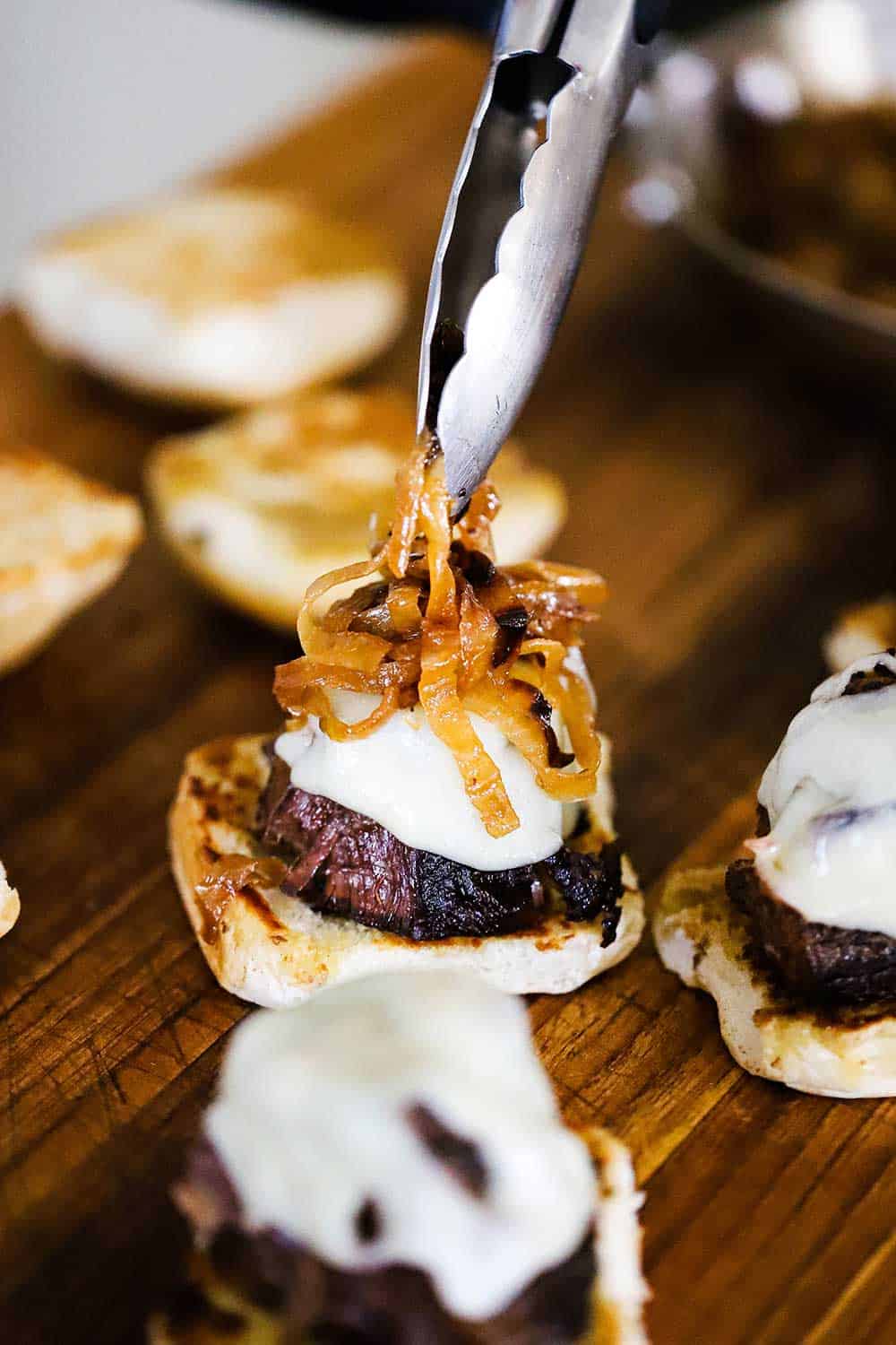 A pair of tongs being used to place a mound of caramelized onions on top of a pot roast with melted cheese sitting on the bottom of a bun. 