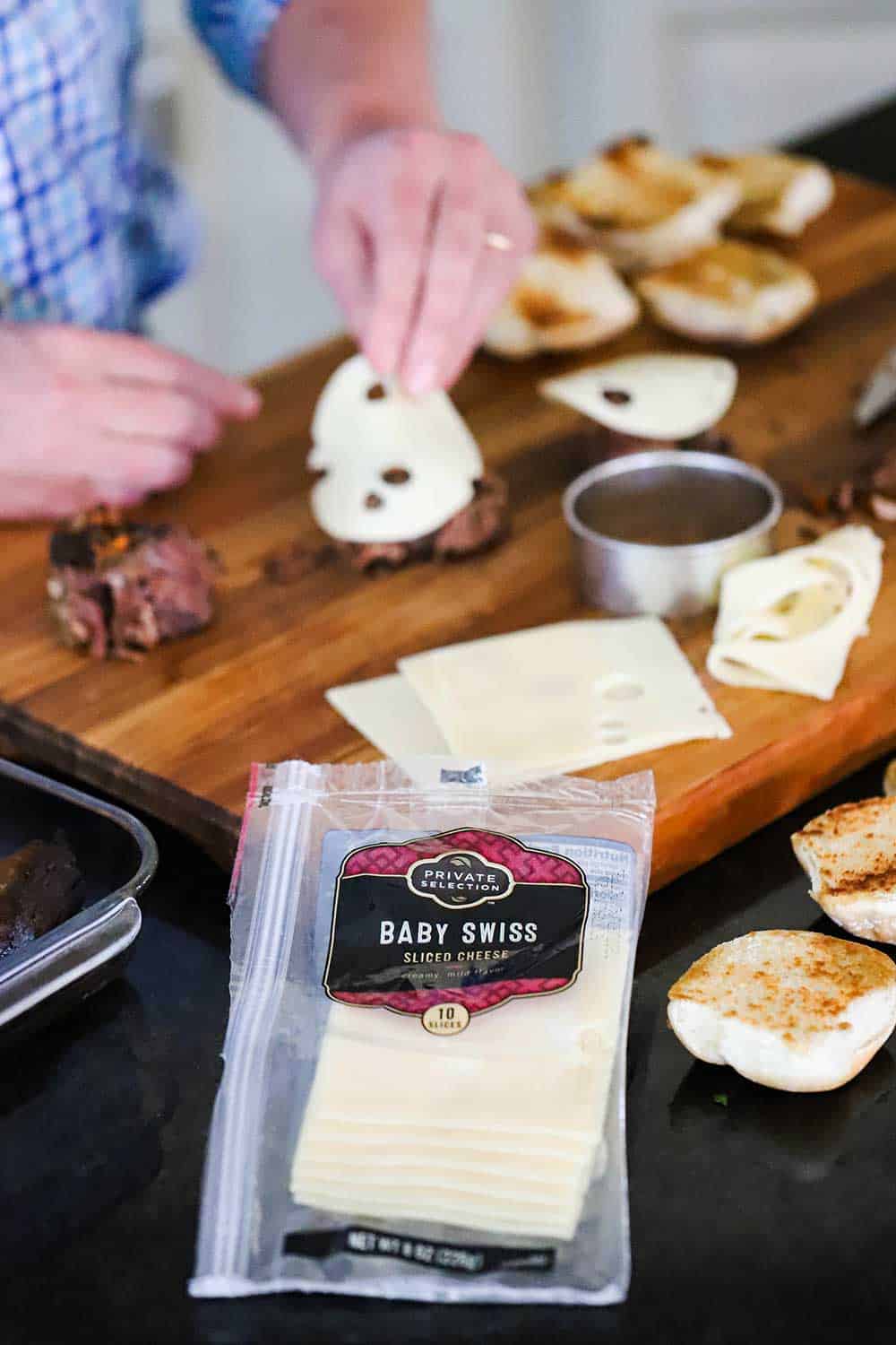 A person adding a circular slice of baby Swiss cheese on top of a piece of pot roast on a bun, surrounded by other cheese slices, cut open dinners rolls, and a cookie cutter. 